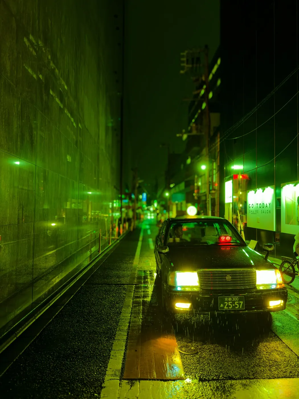 A taxi waits with its headlights on in a narrow Kyoto street at night, rain falling through a green glow from the glass buildings.
